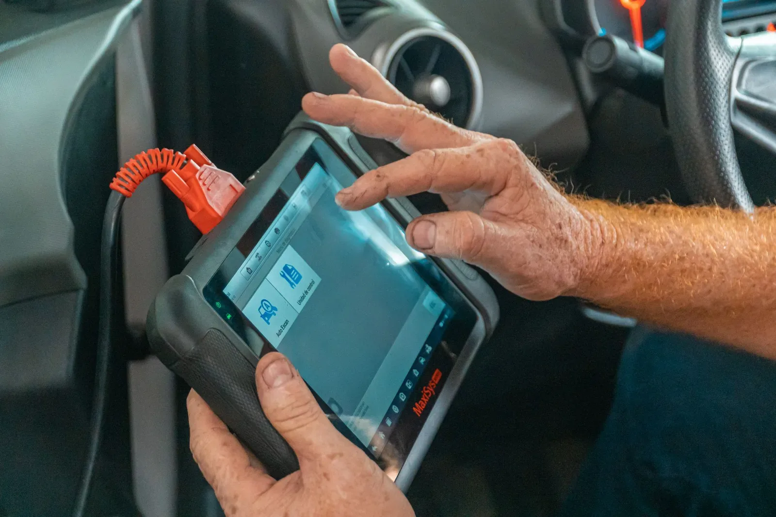 Hand schließt ein OBD-Diagnosegerät an die Fehlerspeicher-Buchse im Fußraum eines Autos an, im Hintergrund unscharf das Cockpit.